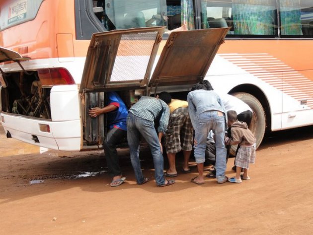 Zeven mensen staan te turen naar de kapotte motor van een bus. 