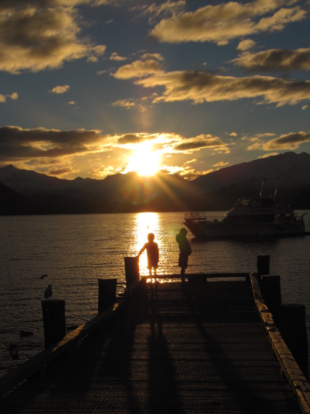 Twee kinderen staan bij Lake Wanaka te kijken hoe de zon achter de besneeuwde bergen verdwijnt